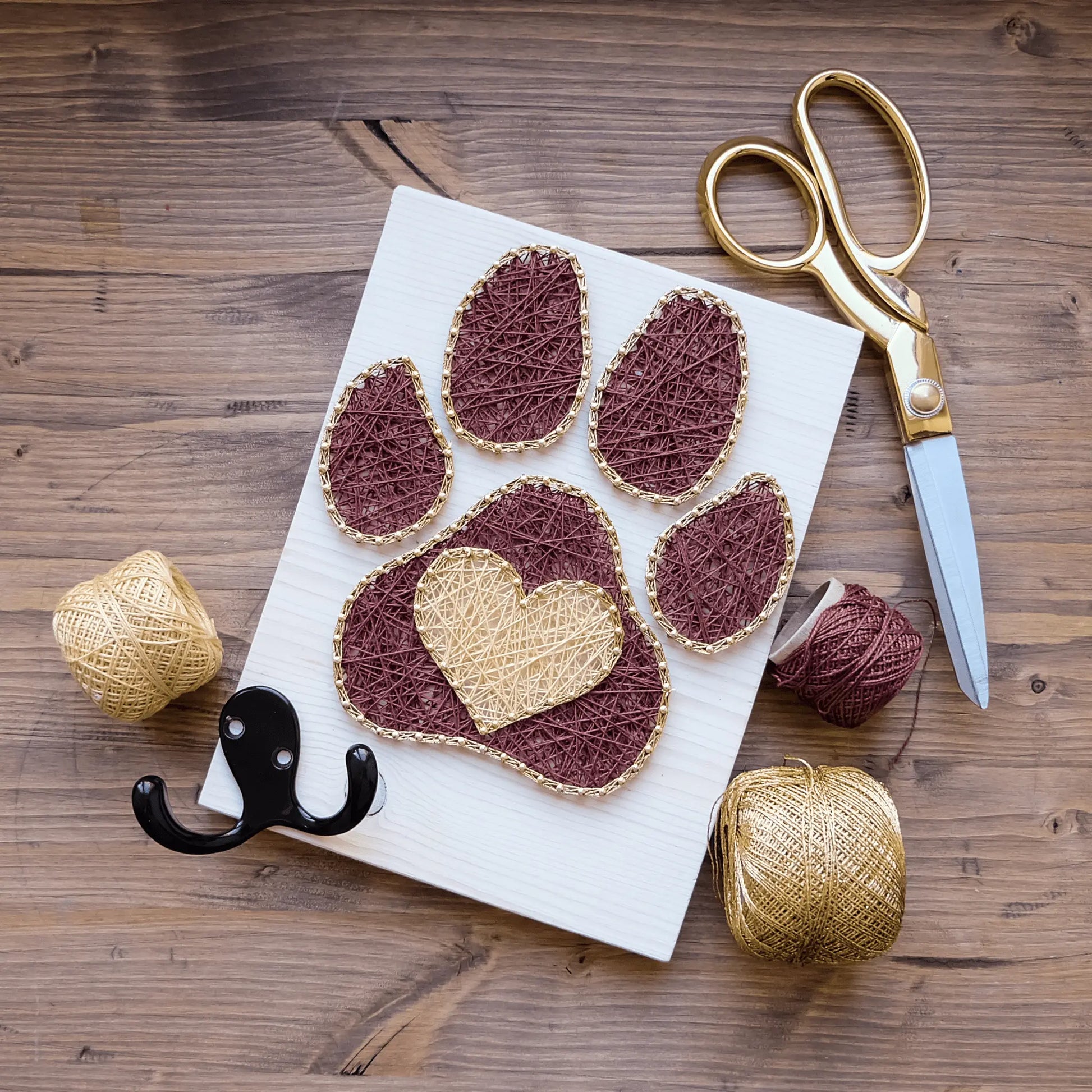 Dog paw string art on a white wooden board with brown and gold threads, paired with crafting tools like scissors and yarn.