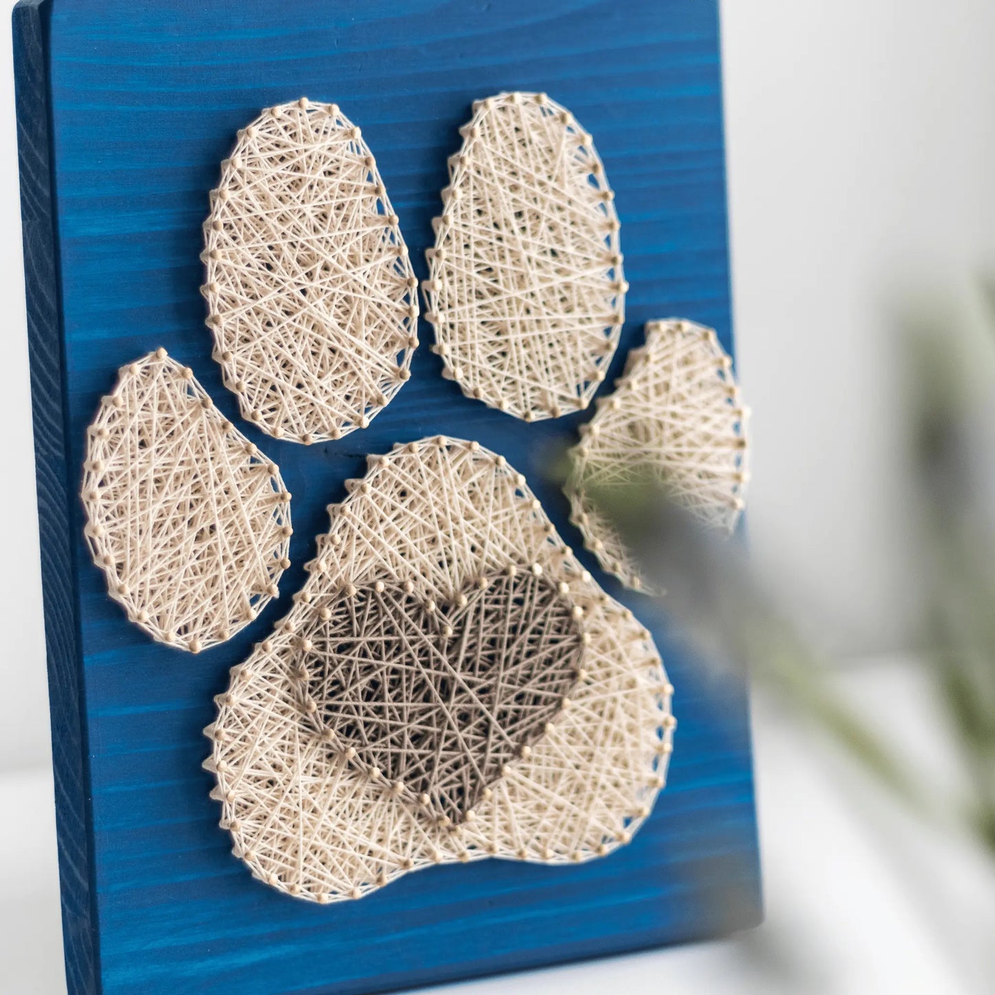 Dog paw string art pattern crafted with natural beige threads on a blue wooden board, emphasizing a heart-shaped center.