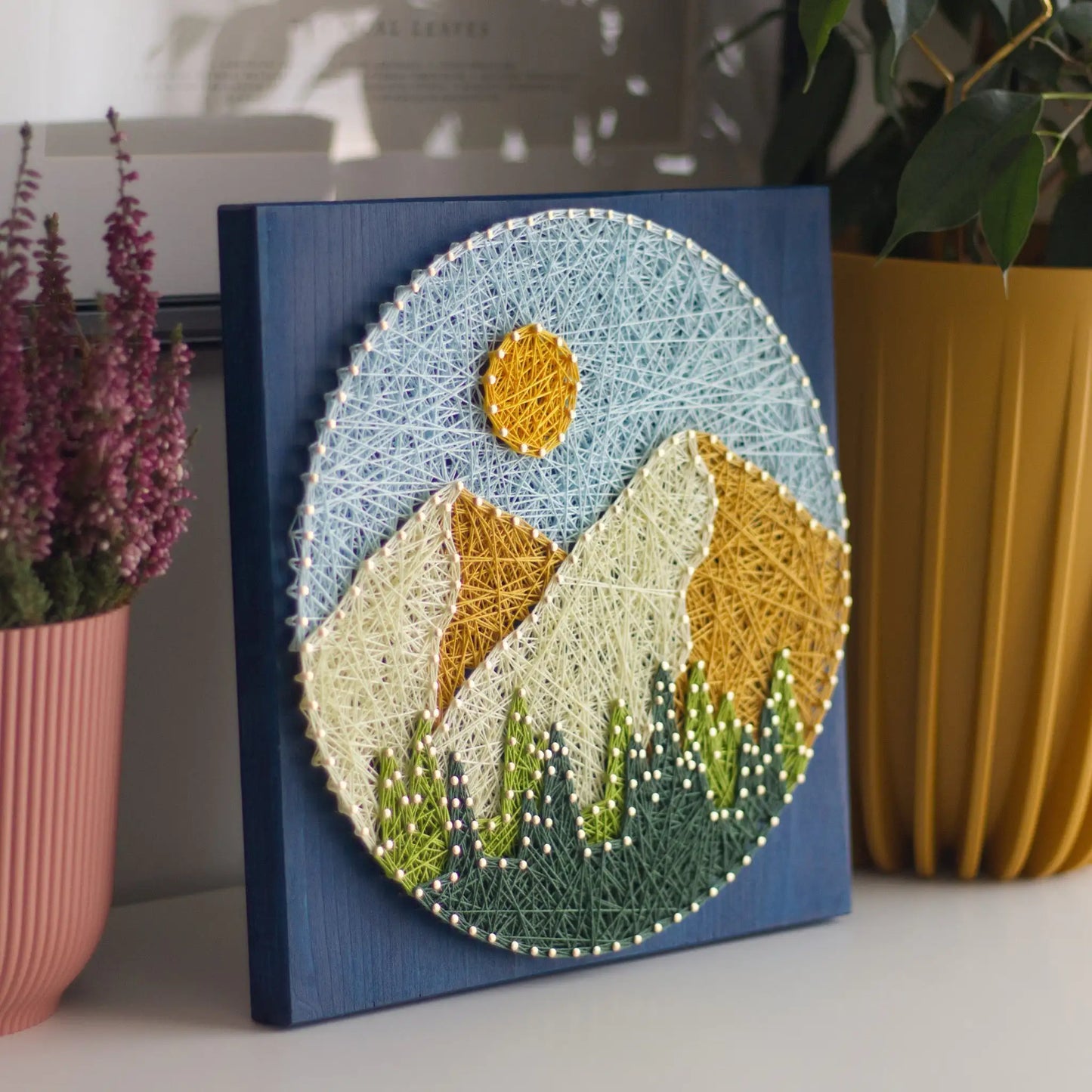 String art of a mountain landscape on a blue background, surrounded by potted plants.
