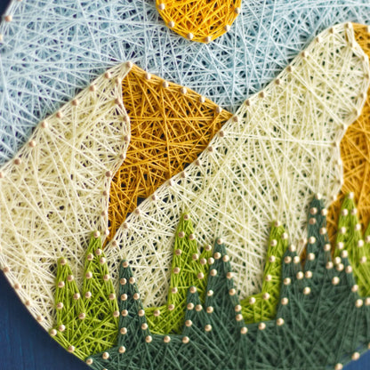 Finished round mountain string art displayed on a shelf, showing layered peaks, yellow sun, and green trees on a blue stained wood base as nature wall decor.