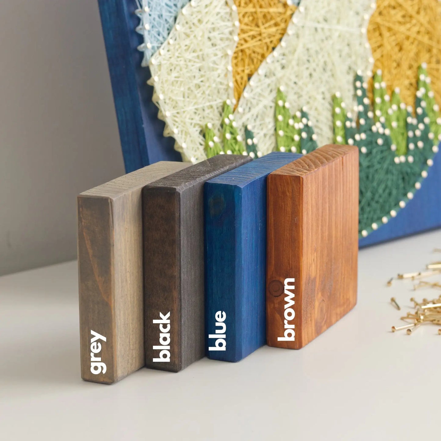 Wood base stain options for the DIY string art kit, showing grey, black, blue, and brown stained square wooden boards displayed side by side.