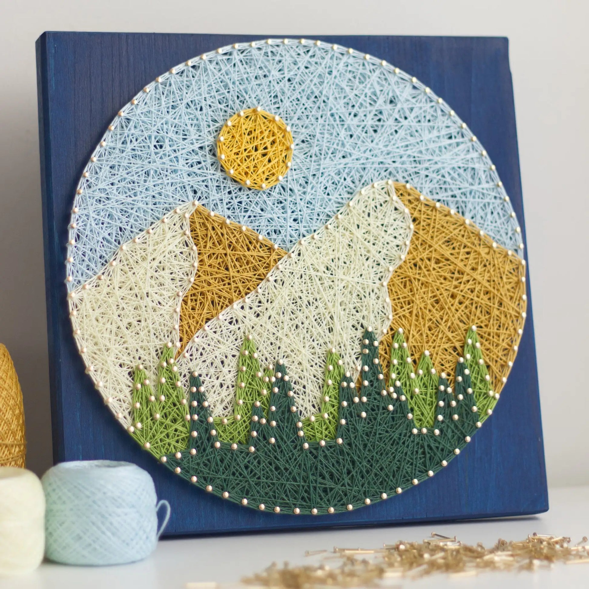 Round mountain string art on a dark blue stained wood base, showing layered cream and golden peaks, a yellow sun, and green trees, with balls of thread and scattered nails in the foreground.