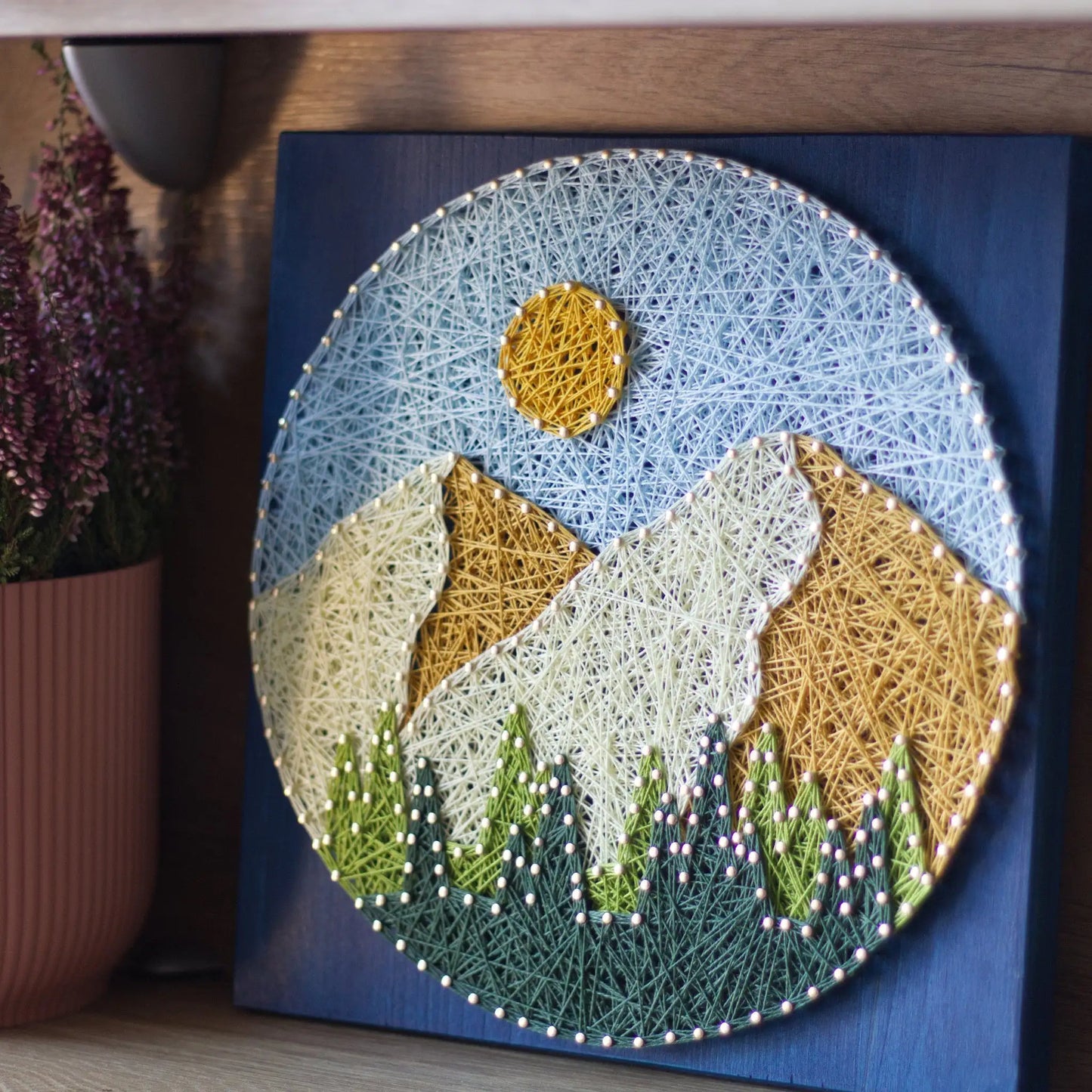 Finished round mountain string art displayed on a shelf, showing layered peaks, yellow sun, and green trees on a blue stained wood base as nature wall decor.