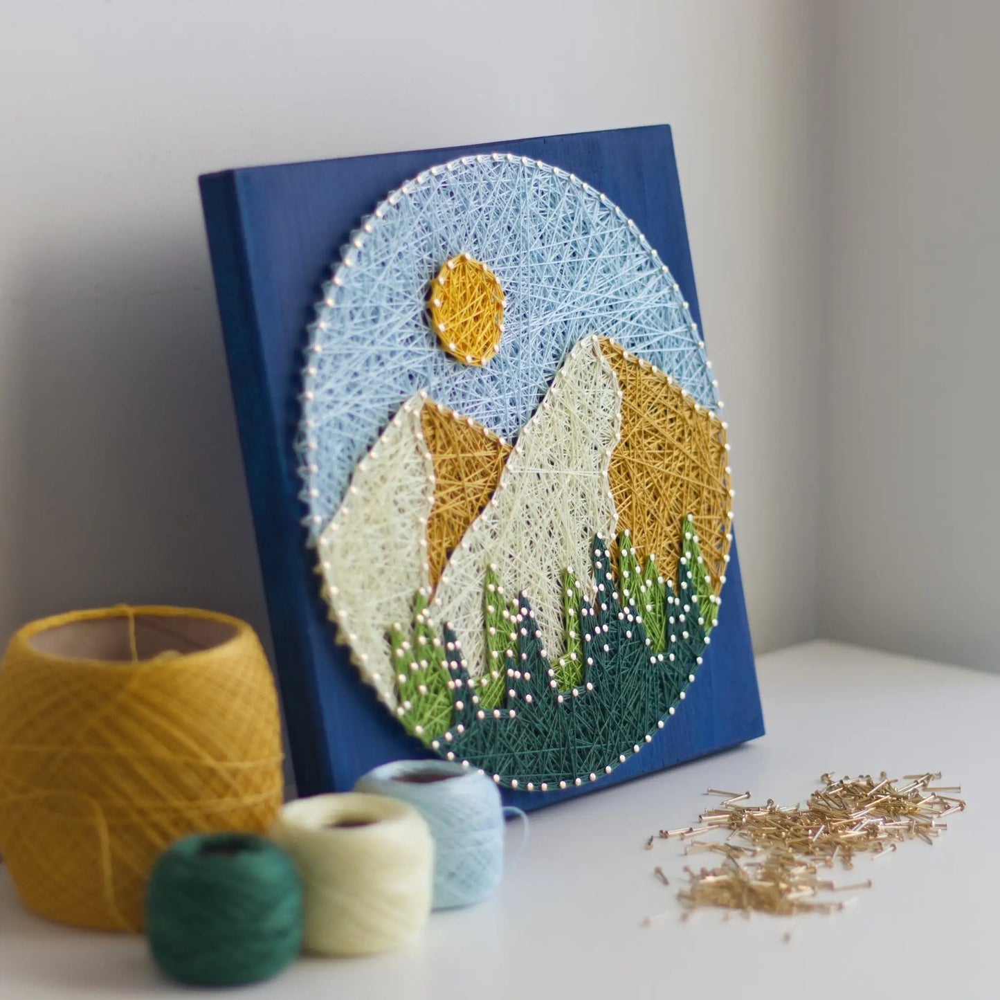 Round mountain string art displayed on a blue stained wood base, styled with yellow and green thread spools and loose nails on a white surface.