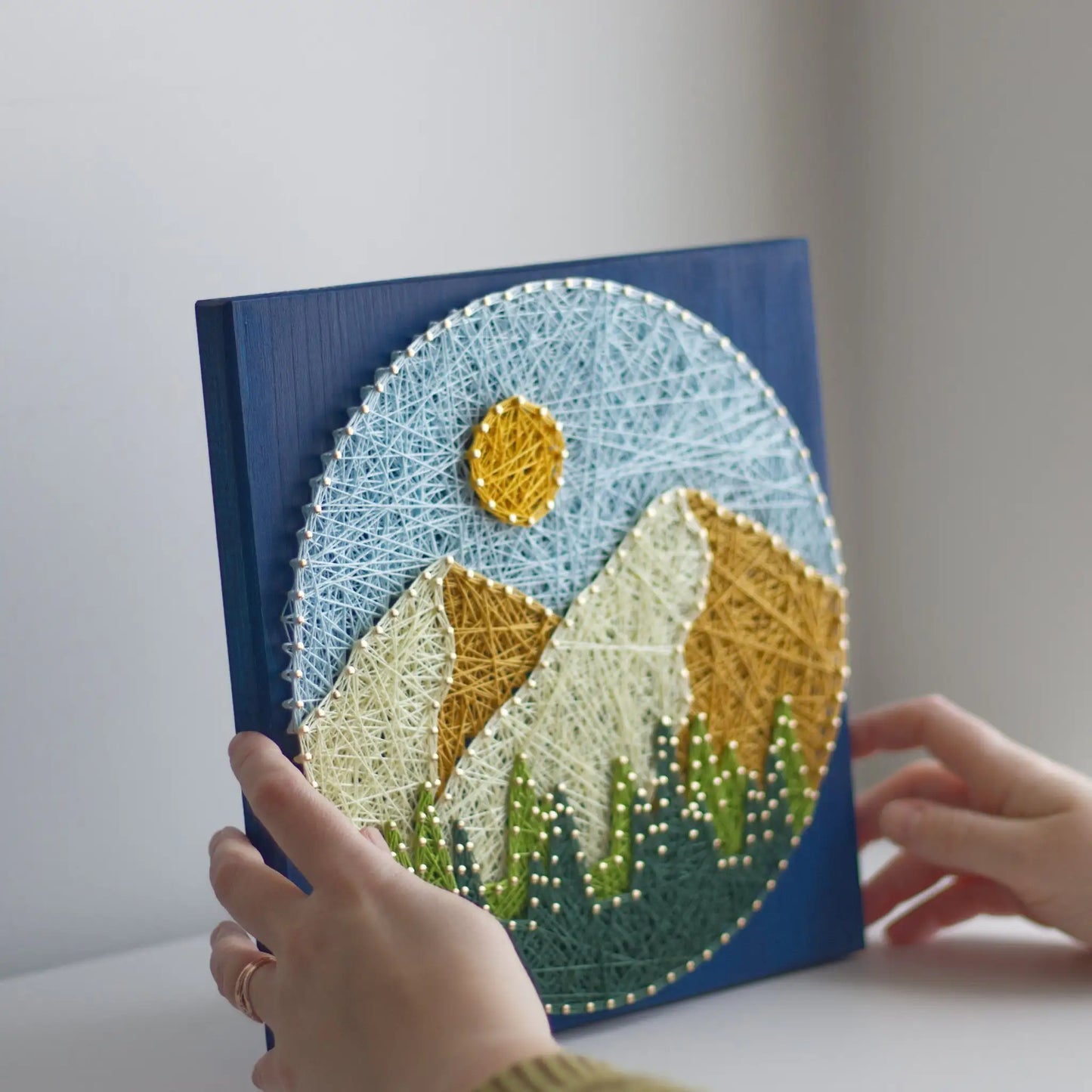 Hands holding a round mountain string art piece on a blue stained wood base, showing layered peaks, sun, and trees in a finished DIY craft.