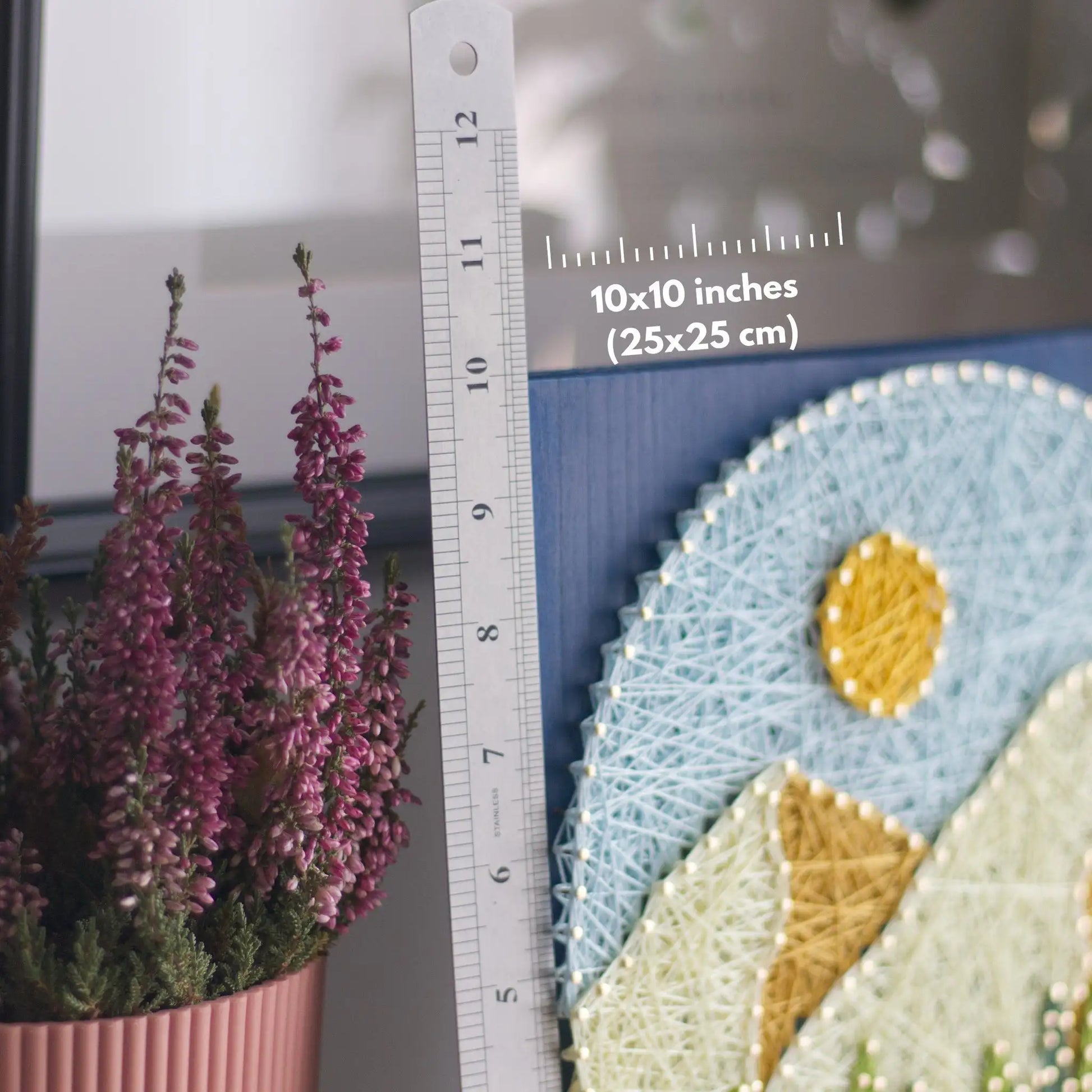 Hands holding a round mountain string art piece on a blue stained wood base, showing layered peaks, sun, and trees in a finished DIY craft.