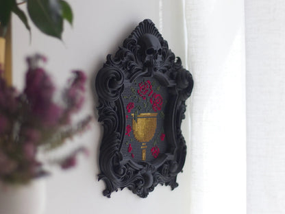 Black gothic skull cross stitch frame hanging on a white wall beside a sheer curtain, seen at an angle, stitched with a gold chalice and red roses.