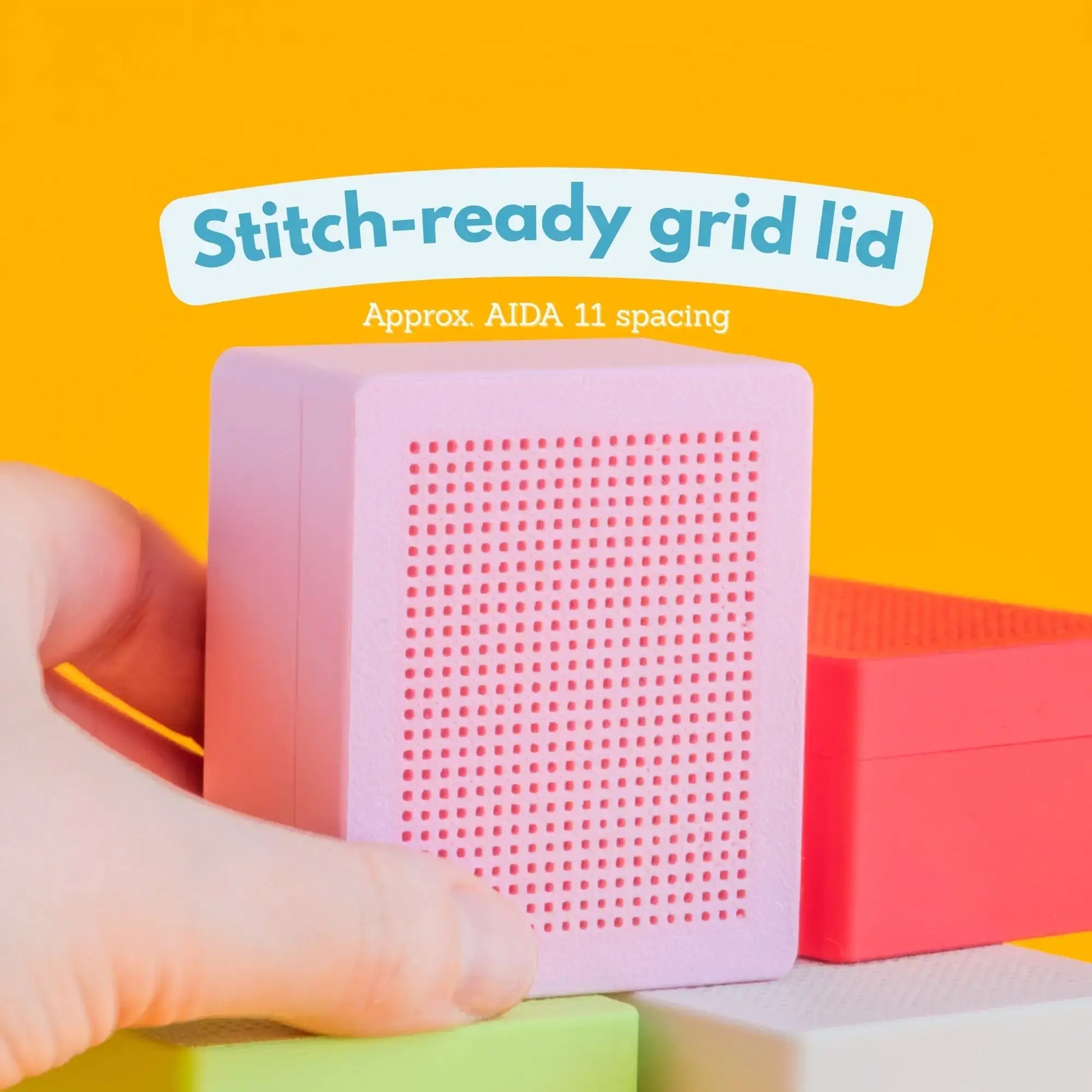 Hand holding a pink 3D-printed cross stitch box, showing the perforated stitch-ready grid lid up close, against a bright yellow background. Text reads Stitch-ready grid lid, approximately Aida 11 spacing.

