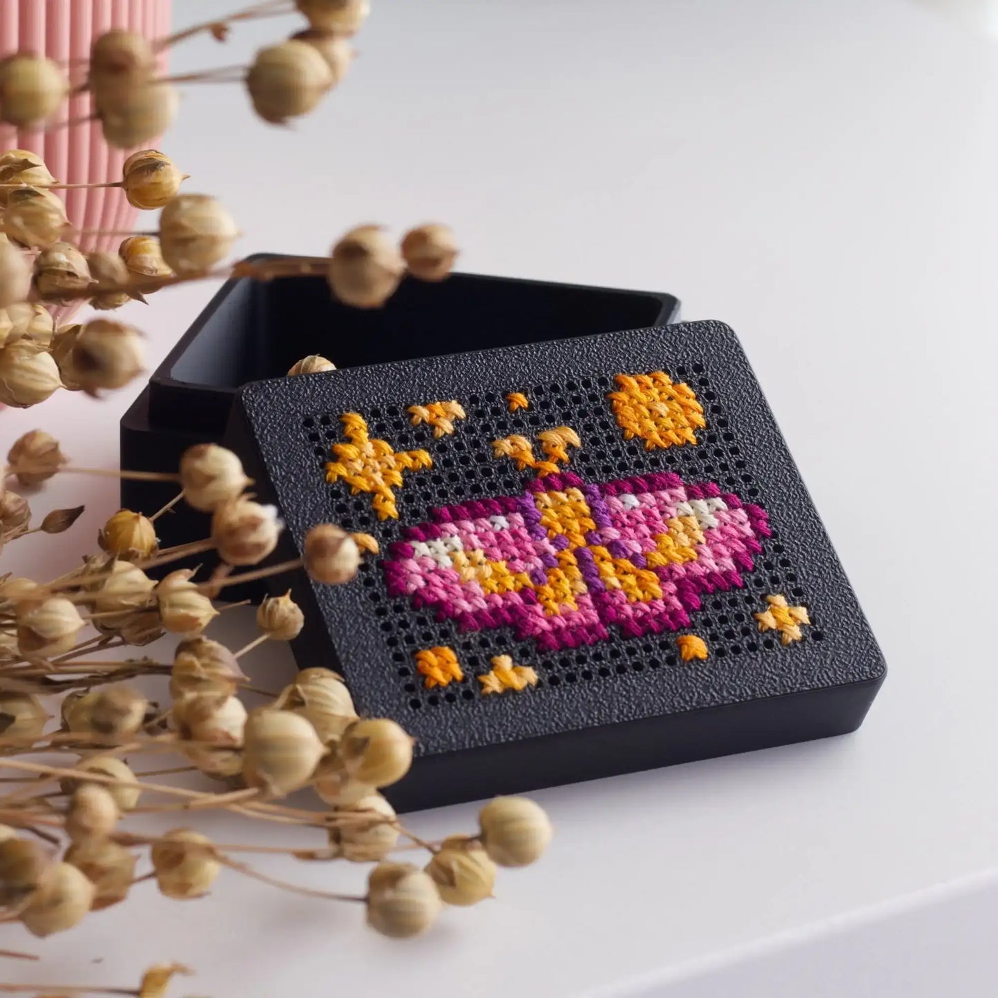 Black 3D-printed cross stitch box with a pink and orange moth design stitched into the perforated lid, shown slightly open on a white surface with dried flowers around it.