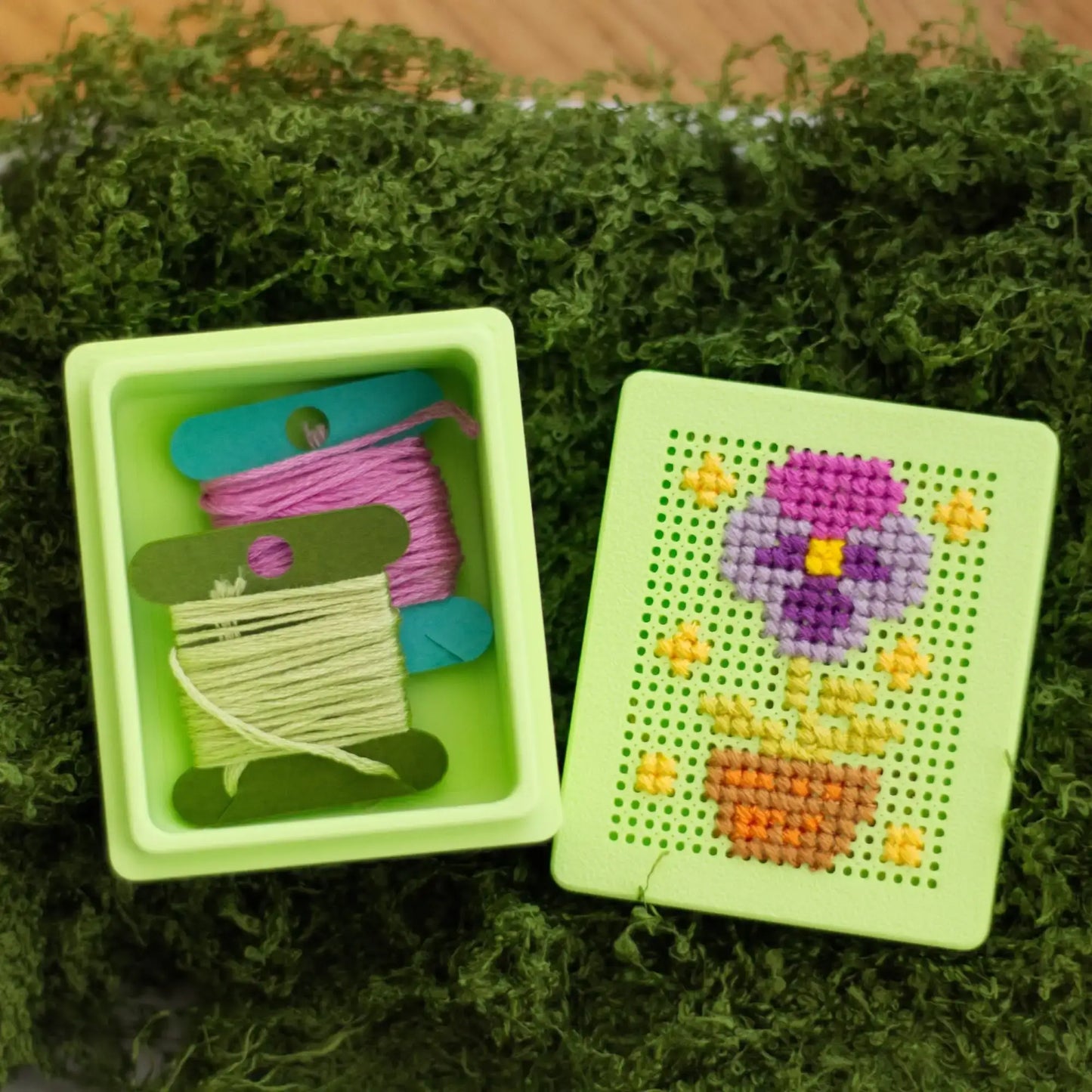 Light green 3D-printed cross stitch box shown open on moss, one part holding embroidery floss and thread spools, the other with a purple flower and pot design stitched into the perforated lid.