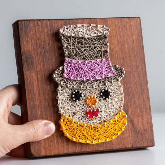 String art snowman on a wooden board held by a hand against a gray background