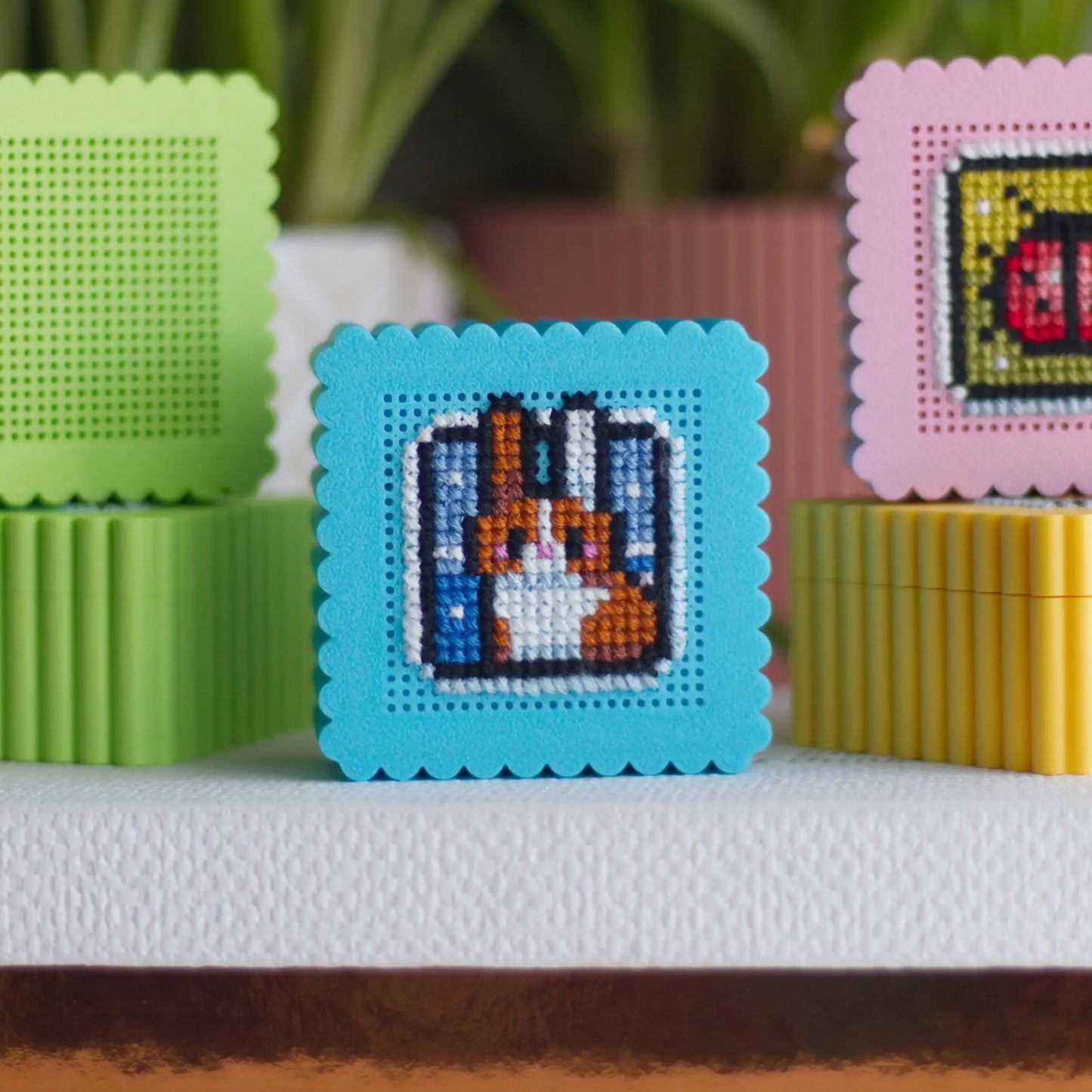 Small embroidery boxes in green, blue, pink, and yellow with perforated lids showing cross stitch bunny and ladybug examples