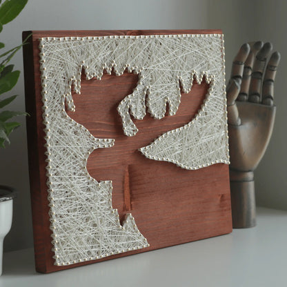 Finished moose silhouette string art made with ivory thread and nails on a stained wooden base, DIY string art kit for adults displayed on a shelf with decor.