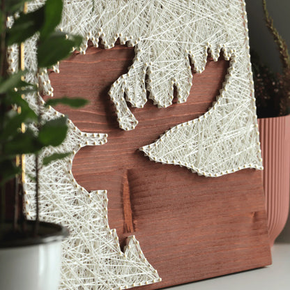 Finished moose silhouette string art made with ivory thread and nails on a stained wooden base, styled with plants to show the DIY string art kit as natural home decor.