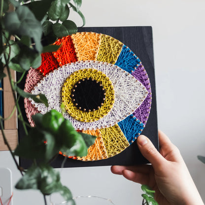 Hand holding a vibrant Third Eye string art design with rainbow colors and a black circular center, surrounded by green foliage for a natural aesthetic.