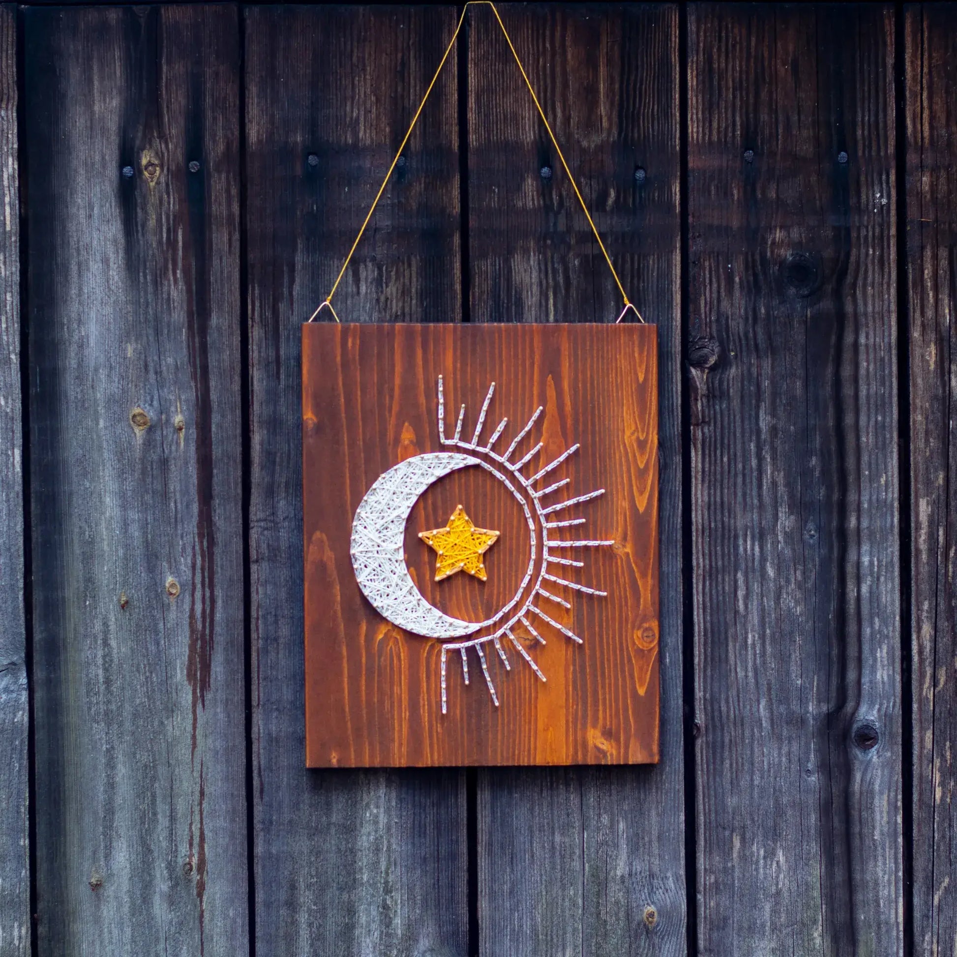 Decorative wooden board featuring a sun, moon, and star string art pattern, hanging against a weathered wooden background.