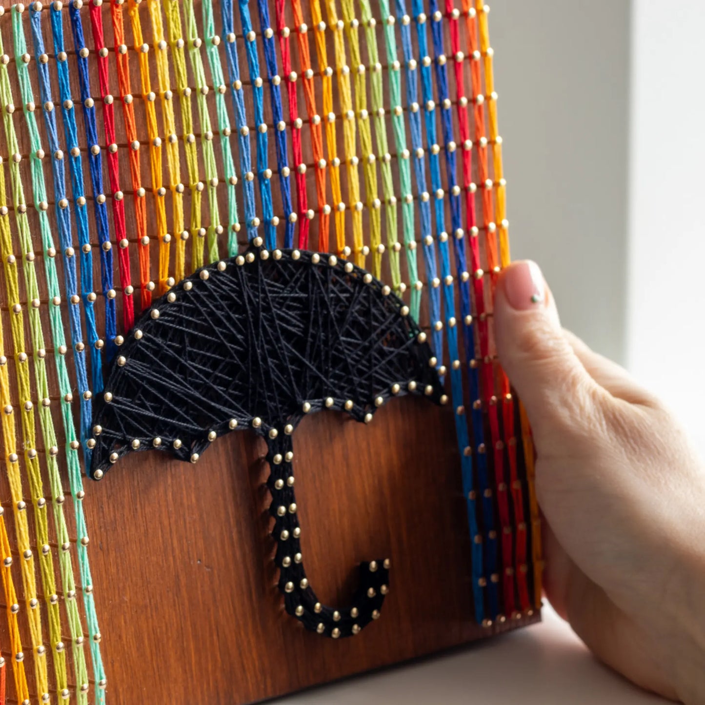 Hand holding a completed string art piece featuring an umbrella and rainbow.