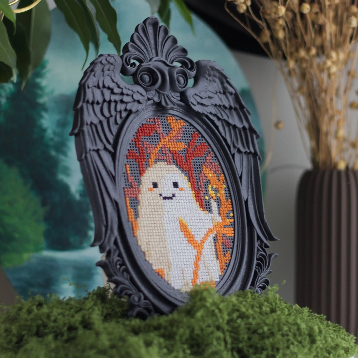Black Victorian angel wing cross stitch frame with completed ghost embroidery displayed on moss base with forest backdrop and dried flowers for whimsigoth gothic home decor