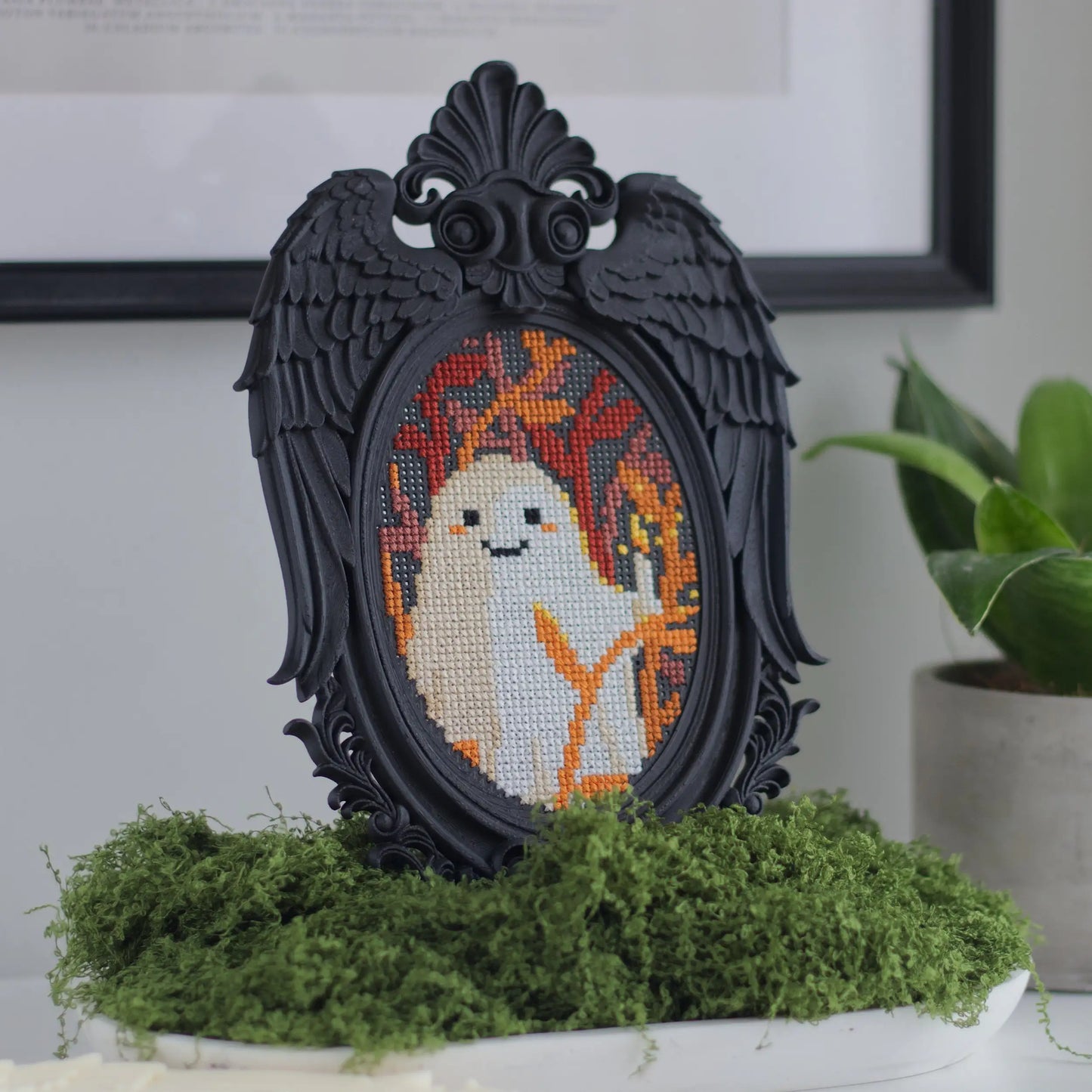Black Victorian angel wing cross stitch frame with ghost embroidery displayed on moss base beside potted plant for whimsigoth gothic home decor styling