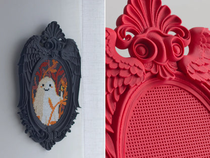Black angel wing cross stitch frame hanging on a wall, and close-up of red frame showing rose and wing details